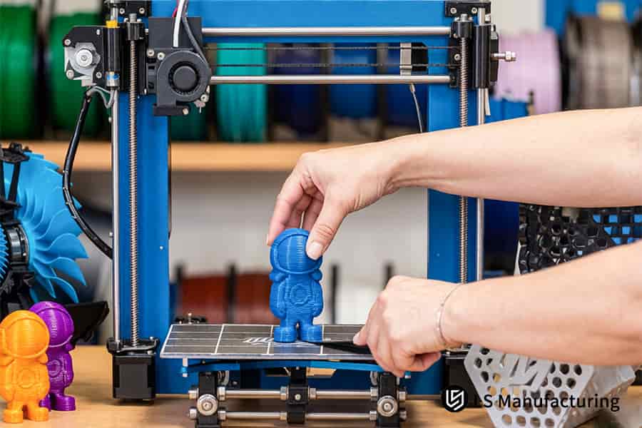 Operator removes blue PLA toy figurine from FDM 3D printer build plate manually.