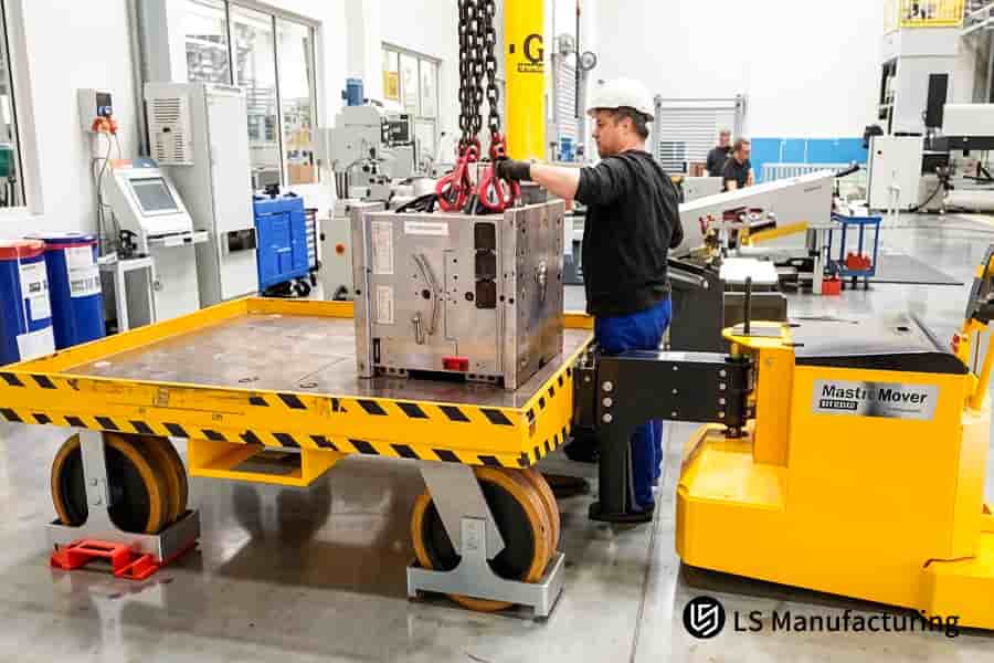 A technician operates a yellow forklift to position a large metal mold component with an overhead chain hoist.